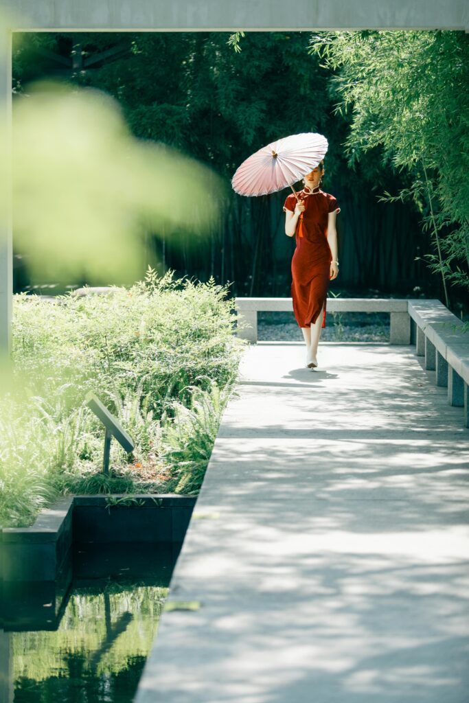 A woman in a red traditional dress walks on a garden path with a parasol.