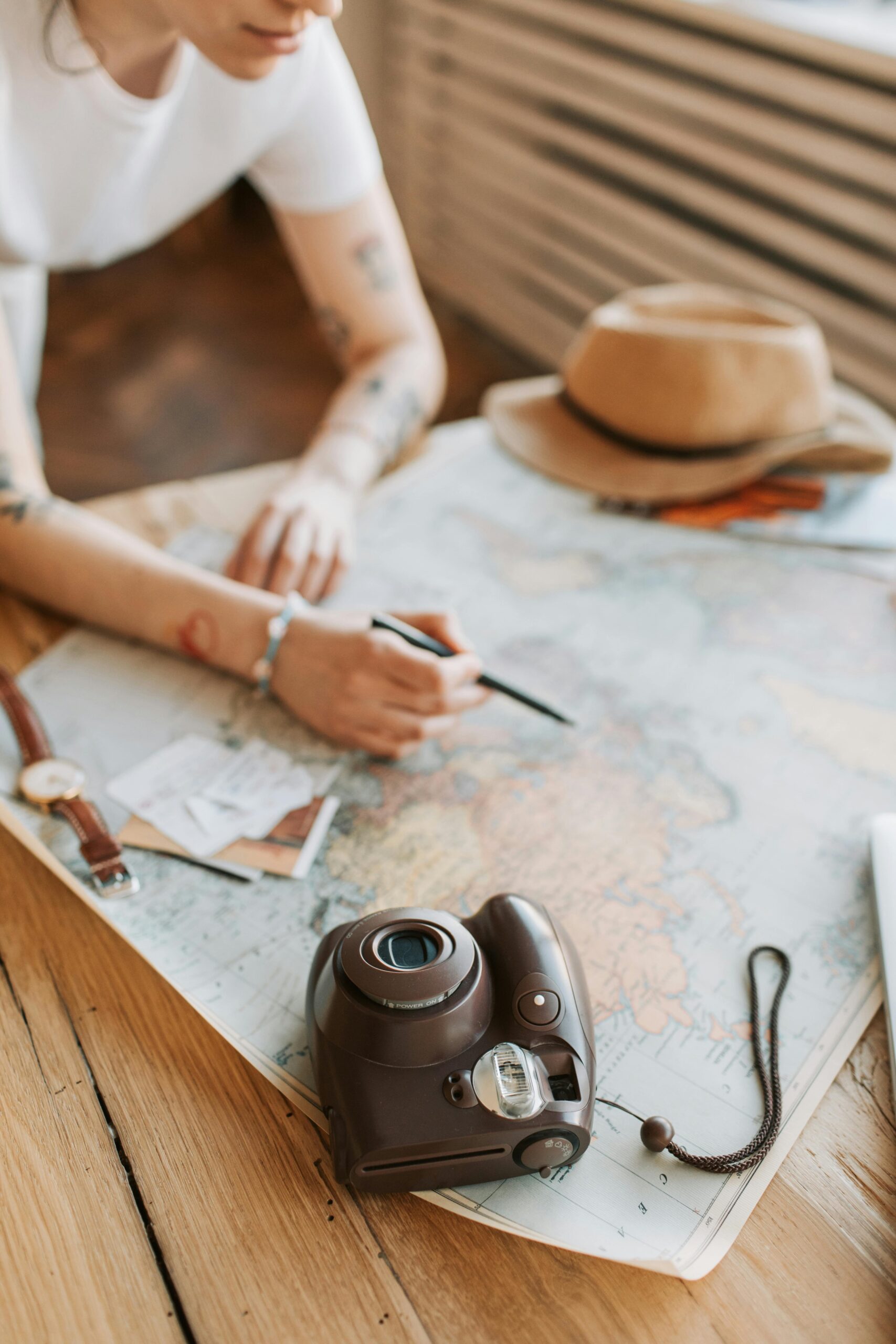 Close-up of a person planning a journey on a world map with a camera and hat on a wooden table.